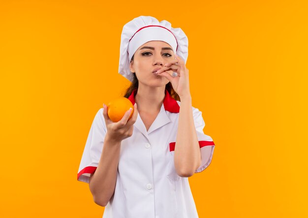 Joven cocinera caucásica complacida en uniforme de chef tiene naranja y gestos signo delicioso delicioso aislado en pared naranja con espacio de copia