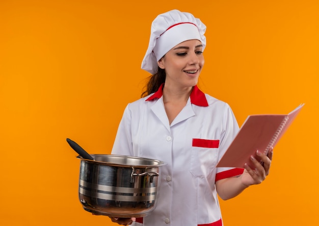 Joven cocinera caucásica complacida en uniforme de chef tiene olla y cuaderno aislado en pared naranja con espacio de copia