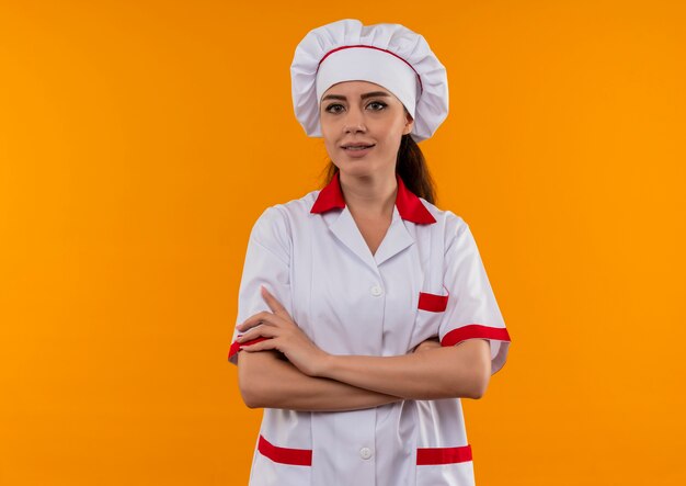 Joven cocinera caucásica confiada en uniforme de chef cruza los brazos y mira a la cámara aislada sobre fondo naranja con espacio de copia