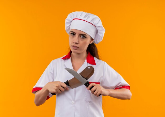 Joven cocinera caucásica confiada en uniforme de chef cruza cuchillos aislados en pared naranja con espacio de copia