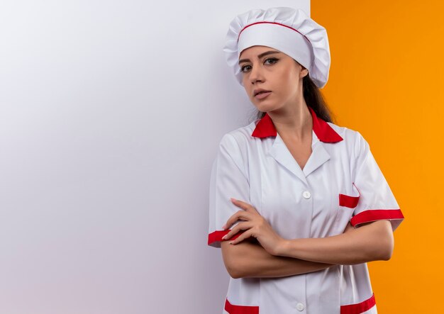 Joven cocinera caucásica confiada en uniforme de chef se encuentra frente a la pared blanca aislada en la pared naranja con espacio de copia