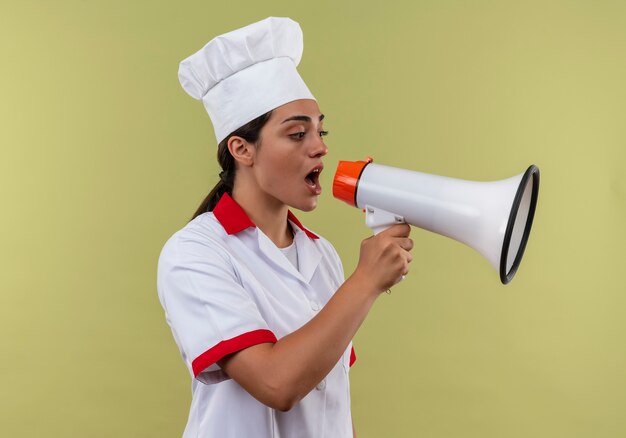 Joven cocinera caucásica confiada en uniforme de chef grita a través del altavoz aislado en la pared verde con espacio de copia