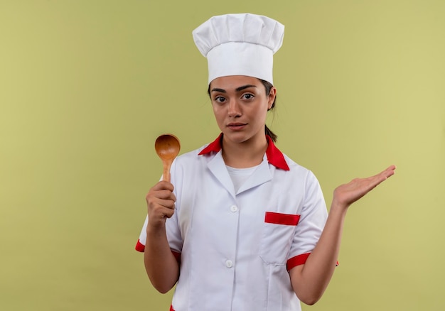 Joven cocinera caucásica confiada en uniforme de chef sostiene una cuchara de madera y mantiene la mano abierta aislada en la pared verde con espacio de copia