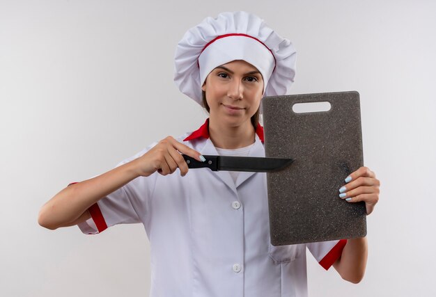 Joven cocinera caucásica confiada en uniforme de chef sostiene tabla de cortar y cuchillo aislado sobre fondo blanco con espacio de copia