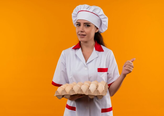 Joven cocinera caucásica confiada en uniforme de chef tiene lote de huevos y pulgares arriba aislados en la pared naranja con espacio de copia