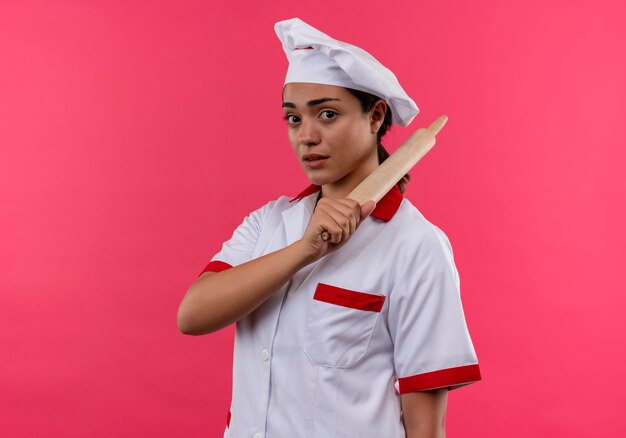 Joven cocinera caucásica confiada en uniforme de chef tiene rodillo aislado en pared rosa con espacio de copia