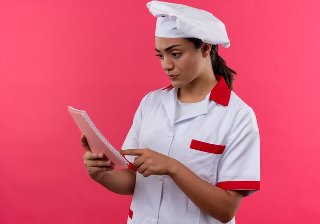 Joven cocinera caucásica confiada en uniforme de chef se ve y apunta al cuaderno aislado en la pared rosa con espacio de copia