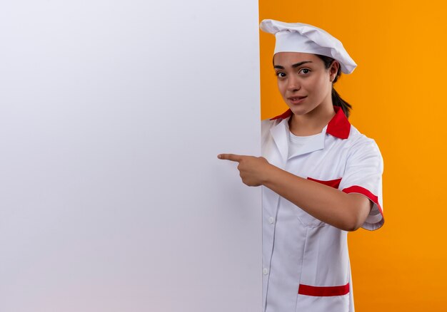 Joven cocinera caucásica molesta en uniforme de chef está detrás de la pared blanca y apunta a la pared aislada sobre fondo naranja con espacio de copia