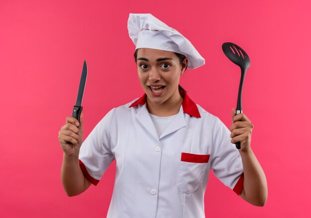 Joven cocinera caucásica molesta en uniforme de chef sostiene cuchillo y espátula aislado sobre fondo rosa con espacio de copia