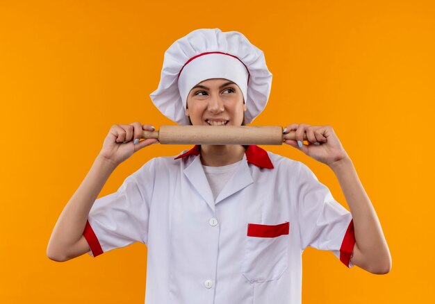 Joven cocinera caucásica molesta en uniforme de chef sostiene y finge morder el rodillo aislado sobre fondo naranja con espacio de copia