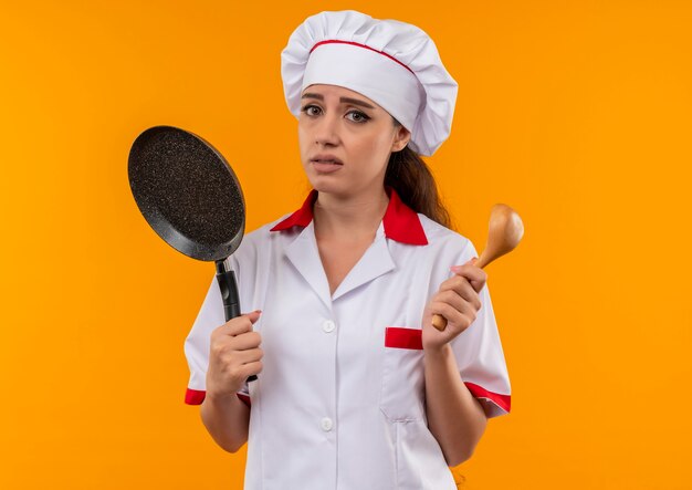 Joven cocinera caucásica molesta en uniforme de chef sostiene una sartén y una cuchara de madera aislada sobre fondo naranja con espacio de copia