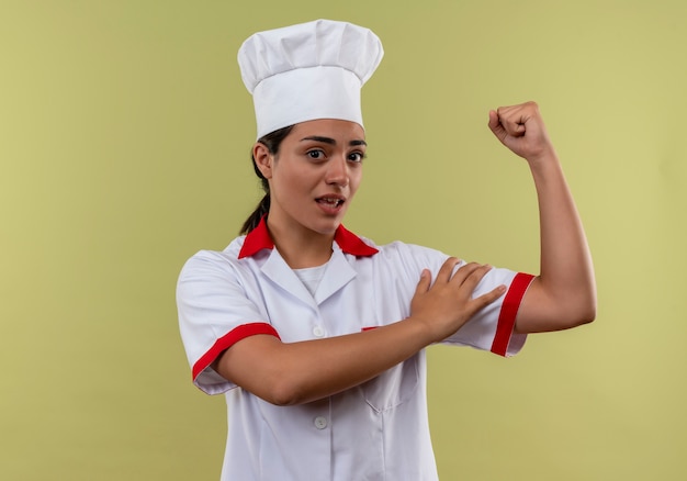 Joven cocinera caucásica molesta en uniforme de chef tensa bíceps y pone mano aislada sobre fondo verde con espacio de copia