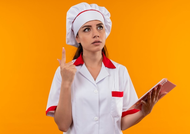 Joven cocinera caucásica pensativa en uniforme de chef sostiene el cuaderno y apunta hacia arriba aislado en la pared naranja con espacio de copia