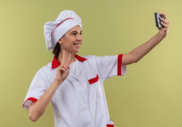 Joven cocinera caucásica sonriente en uniforme de chef mira el teléfono y gestos signo de mano de victoria en verde con espacio de copia