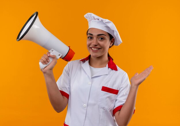 Joven cocinera caucásica sonriente en uniforme de chef sostiene altavoz y mantiene la mano levantada aislada en la pared naranja