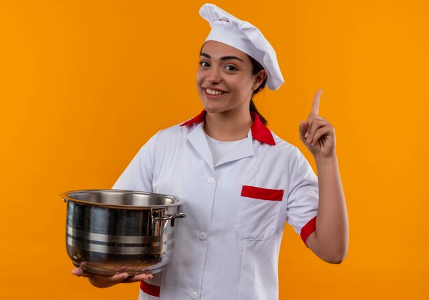 Joven cocinera caucásica sonriente en uniforme de chef sostiene olla y apunta hacia arriba aislado en la pared naranja