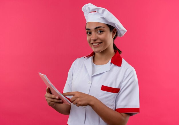Joven cocinera caucásica sonriente en uniforme de chef sostiene portátil y señala con el dedo aislado en la pared rosa con espacio de copia