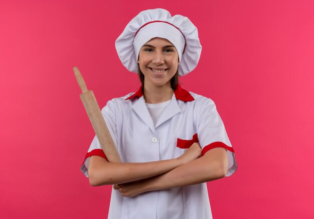 Joven cocinera caucásica sonriente en uniforme de chef sostiene un rodillo con los brazos cruzados aislado en el espacio rosa con espacio de copia