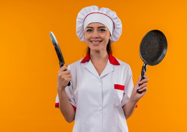 Joven cocinera caucásica sonriente en uniforme de chef sostiene una sartén y un cuchillo aislado en la pared naranja con espacio de copia