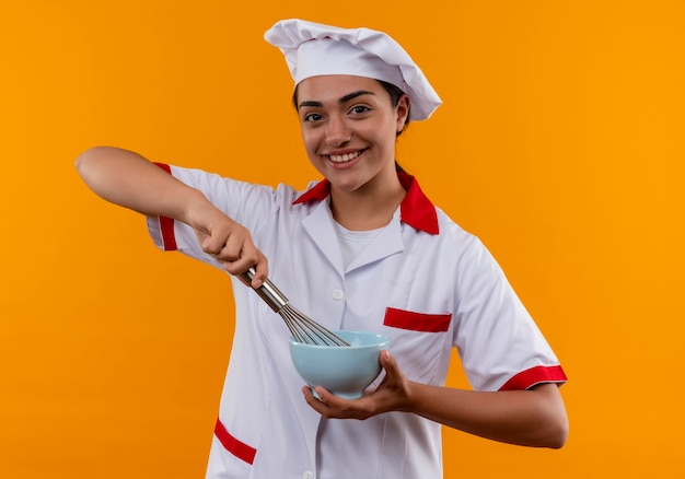 Joven cocinera caucásica sonriente en uniforme de chef sostiene un tazón y un batidor aislado en la pared naranja con espacio de copia