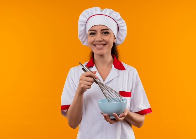 Joven cocinera caucásica sonriente en uniforme de chef sostiene un tazón y un batidor aislado en la pared naranja con espacio de copia