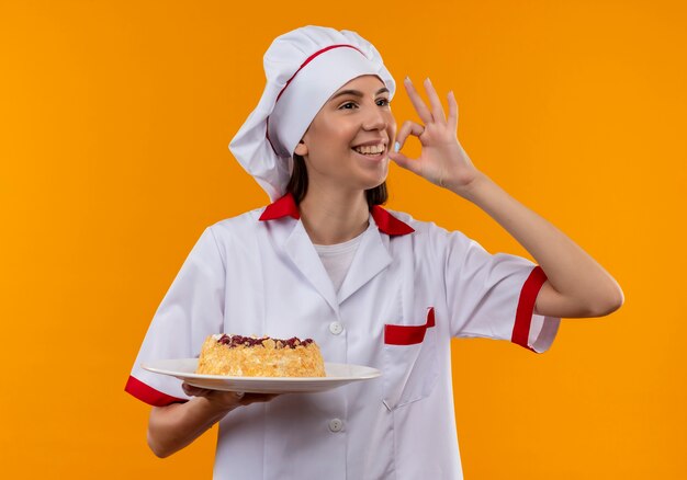 Joven cocinera caucásica sonriente en uniforme de chef sostiene la torta en el plato y gesticula un delicioso cartel aislado en el espacio naranja con espacio de copia