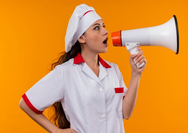 Joven cocinera caucásica sorprendida en uniforme de chef grita a través del altavoz aislado en la pared naranja con espacio de copia