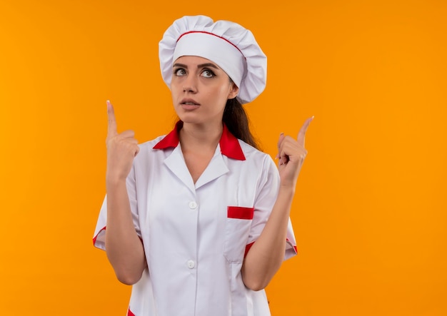 Joven cocinera caucásica sorprendida en uniforme de chef mira y apunta hacia arriba aislado en la pared naranja con espacio de copia