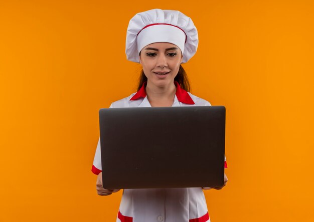 Joven cocinera caucásica sorprendida en uniforme de chef sostiene y mira portátil aislado en pared naranja con espacio de copia