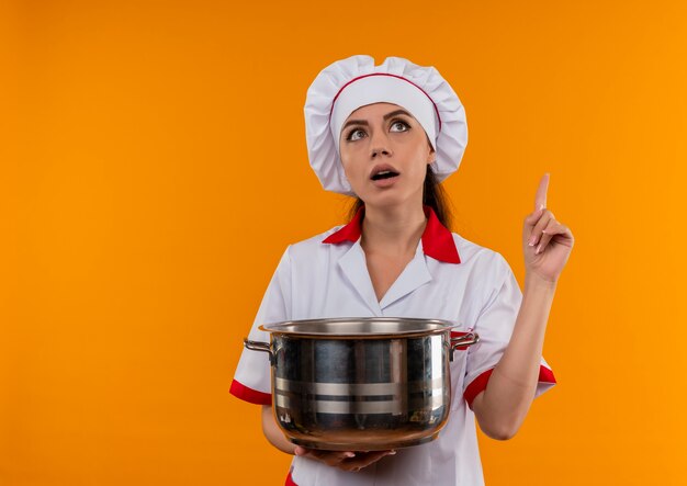 Joven cocinera caucásica sorprendida en uniforme de chef sostiene la olla y apunta hacia arriba aislado en la pared naranja con espacio de copia