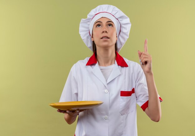 Foto gratuita joven cocinera caucásica sorprendida en uniforme de chef sostiene placa y apunta hacia arriba aislado sobre fondo verde con espacio de copia