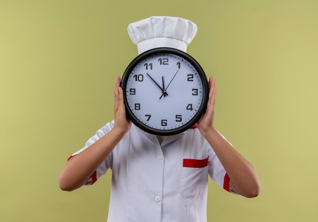 Joven cocinera caucásica en uniforme de chef cierra la cara woth reloj aislado sobre fondo verde con espacio de copia