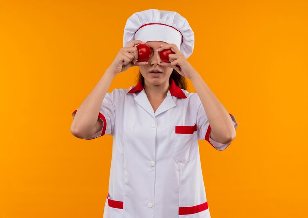 Joven cocinera caucásica en uniforme de chef cierra los ojos con tomates aislados sobre fondo naranja con espacio de copia