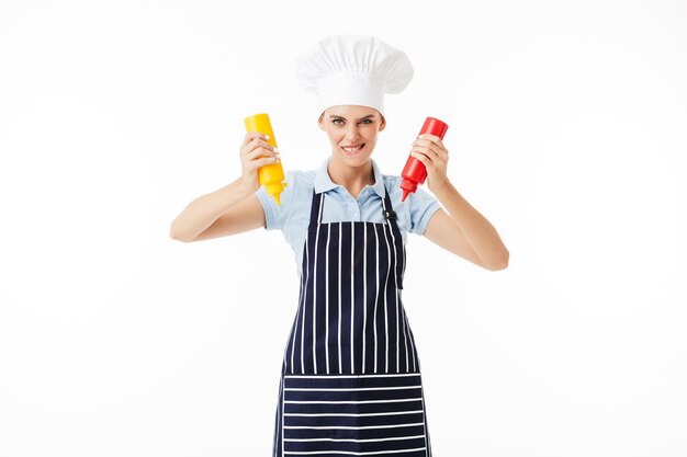 Joven cocinera con delantal a rayas y sombrero blanco mirando juguetonamente a la cámara mientras sostiene botellas de ketchup y mostaza en las manos sobre fondo blanco.