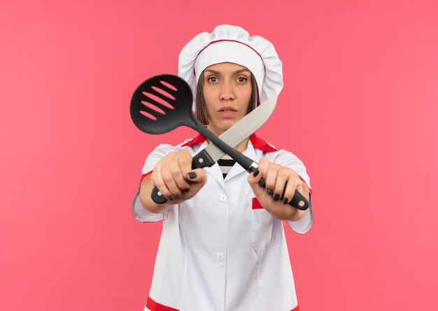 Joven cocinera disgustada en uniforme de chef sosteniendo una espátula y un cuchillo y gesticulando no con ellos a la cámara aislada sobre fondo rosa con espacio de copia