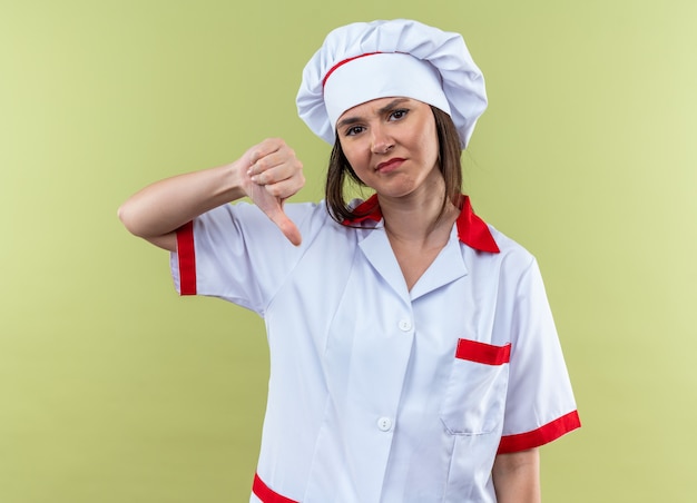 Foto gratuita joven cocinera disgustada vistiendo uniforme de chef mostrando el pulgar hacia abajo aislado en la pared verde oliva