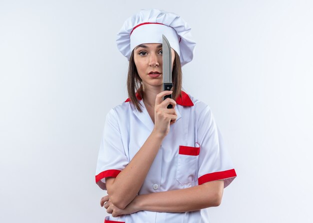 Joven cocinera disgustada vistiendo uniforme de chef ojo cubierto con cuchillo aislado sobre fondo blanco.