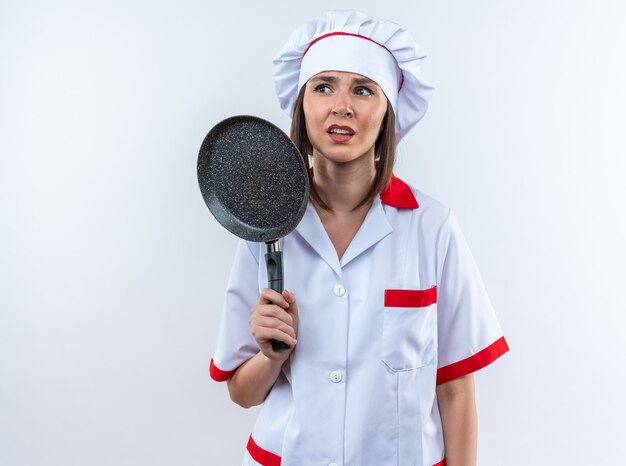 Joven cocinera disgustada vistiendo uniforme de chef sosteniendo una sartén aislado sobre fondo blanco.