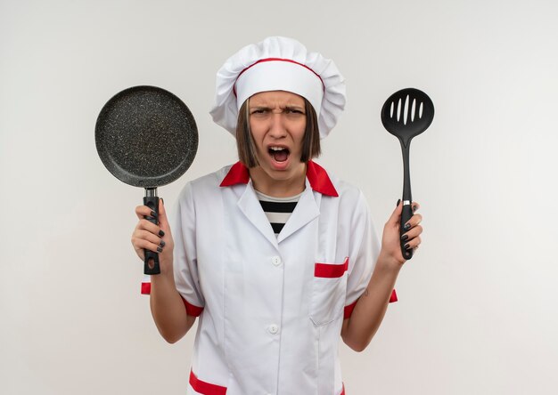 Joven cocinera enojada en uniforme de chef sosteniendo una espátula y una sartén mirando aislado en blanco