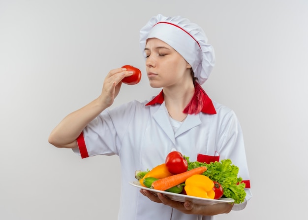 Joven cocinera rubia complacida en uniforme de chef sostiene verduras en un plato y finge oler tomate aislado en la pared blanca
