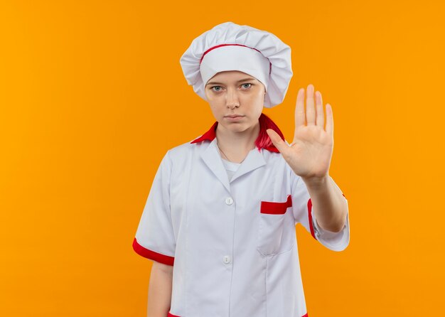 Joven cocinera rubia confiada en gestos de uniforme de chef se detiene aislado en la pared naranja