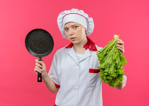 Joven cocinera rubia sorprendida en uniforme de chef sostiene una sartén y ensalada aislada en la pared rosa