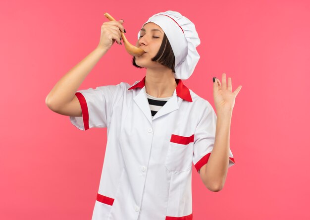 Joven cocinera en uniforme de chef comiendo de una cuchara haciendo bien firmar con los ojos cerrados aislado sobre fondo de color rosa