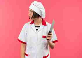 Foto gratuita joven cocinera en uniforme de chef sosteniendo un cuchillo y mirando al lado aislado sobre fondo rosa con espacio de copia