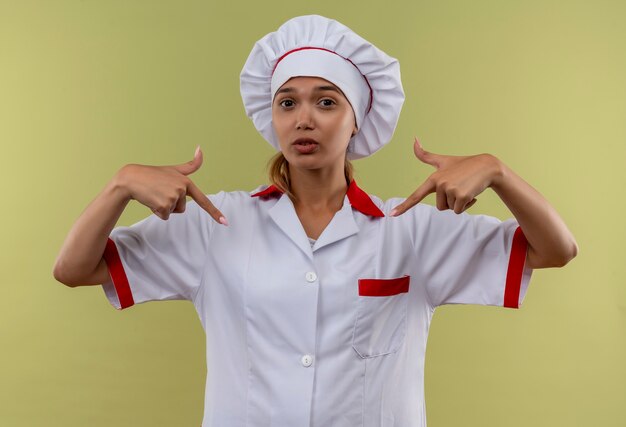 Joven cocinera vestida con uniforme de chef apunta a sí misma en la pared verde aislada