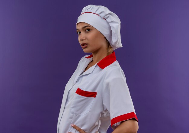 Foto gratuita joven cocinera vestida con uniforme de chef puso sus manos en la cadera en la pared aislada con espacio de copia