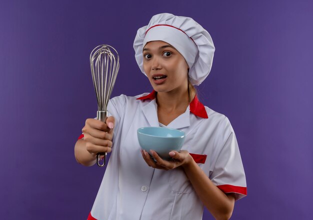 Joven cocinera vestida con uniforme de chef sosteniendo un batidor y un tazón en la pared aislada