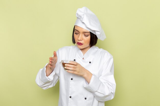 Una joven cocinera de vista frontal en traje de cocinero blanco y gorra sosteniendo una taza de café en polvo en la pared verde