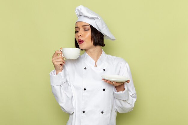 Una joven cocinera de vista frontal en traje de cocinero blanco y gorra sosteniendo la taza en la pared verde