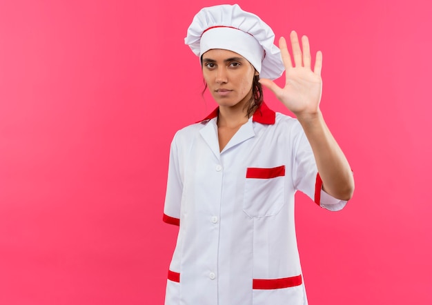 Joven cocinera vistiendo uniforme de chef mostrando gesto de parada en la pared rosa aislada con espacio de copia